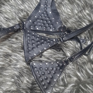 Könnte beinhalten: Schwarzes Ledergeschirr mit silberfarbenen Nieten. Das Geschirr hat eine dreieckige Ober- und Unterseite, die durch Riemen und Metallringe verbunden sind. Das Leder hat eine strukturierte, genoppte Oberfläche.