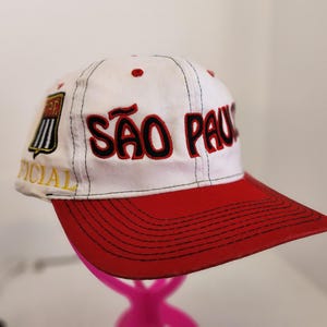 May include: White and red baseball cap with the text "SÃO PAULO" in red and black. The cap has a red brim and a small patch on the side with the word "OFFICIAL".