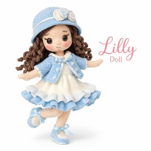 Op de afbeelding: Een handgemaakte gehaakte pop genaamd "Lilly" met een lichtblauwe hoed, jas en schoenen, en een witte jurk. De pop heeft bruin krullend haar en roze wangen. De tekst "Lilly Doll" is in roze geschreven.