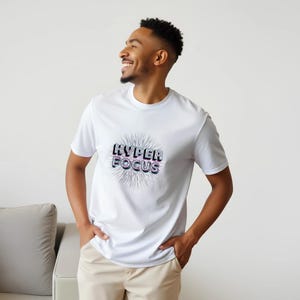 Puede incluir: Una camiseta blanca de manga corta con el texto "HYPER FOCUS" impreso en un estilo de fuente audaz, superpuesto y ligeramente distorsionado. El texto está centrado en el pecho y rodeado por un estallido radiante de líneas finas y oscuras.