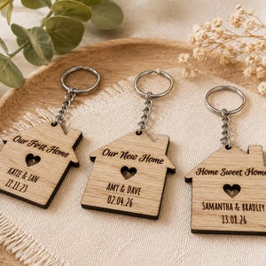 May include: Three wooden house keyrings with silver chains. Each features a heart-shaped cutout and engraved text: "Our First Home," "Our New Home," and "Home Sweet Home." Names and dates are also engraved on each keyring.