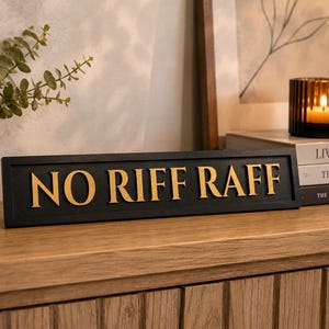 May include: A black wooden sign with the words "NO RIFF RAFF" in gold lettering. The sign is placed on a wooden surface. A candle in a glass holder and books are in the background. A potted plant is visible on the left.