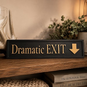 May include: A black rectangular sign with the words "Dramatic EXIT" in gold lettering and a gold arrow pointing down. The sign is displayed on a wooden shelf, with decorative items in the background.