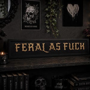 May include: A rectangular black wooden sign featuring the text FERAL AS FUCK in raised, metallic gold-toned lettering with a gothic-inspired font. The sign is positioned horizontally, showcasing a clean, bold design with high-contrast typography.