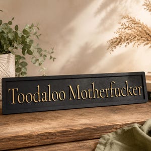 May include: A black wooden sign with the words "Toodaloo Motherfucker" in gold lettering. The sign is rectangular and sits on a wooden surface. Decorative plants and books are in the background.