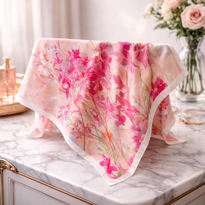 May include: A square scarf with a floral pattern and a white border. The scarf displays a watercolour design of pink and magenta flowers on a light peach background. The scarf is draped over an unseen object.