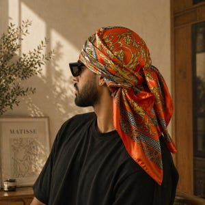 Puede incluir: Un pañuelo de seda naranja con un estampado barroco dorado está atado alrededor de la cabeza. El pañuelo se lleva sobre una camiseta negra. La persona lleva gafas de sol negras.