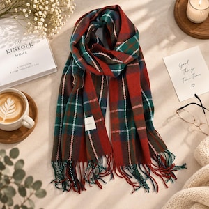 May include: A rectangular scarf featuring a plaid pattern in shades of deep red, forest green, and navy blue with thin white accent lines. The scarf has a soft texture and is finished with long, loose fringe tassels along the bottom edges.
