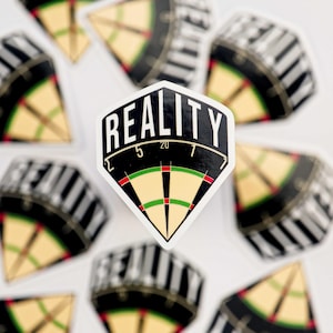 Op de afbeelding: Witte sticker met het woord "REALITY" in dikke, zwarte letters. De sticker heeft de vorm van een dartbord, met een zwart-witte rand en een beige en groen midden. Er liggen meerdere andere dartbordstickers verspreid.