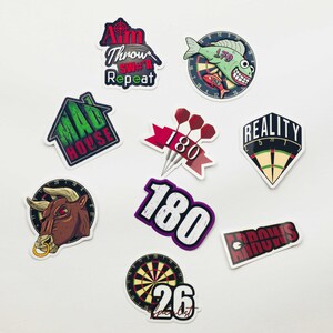 Op de afbeelding: Een verzameling van negen vinylstickers met dart-thema's. De afbeeldingen bevatten een stier, een dartbord met een vis, de tekst Aim Throw Sw#r Repeat, Mad House, drie darts met het getal 180, een Reality-bord en het getal 26.