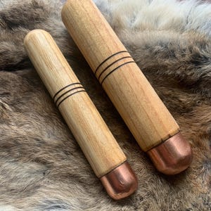 May include: Two cylindrical wooden tools with light tan grain and smooth surfaces. Each tool features three thin, dark, horizontal engraved lines near the centre and a rounded, polished copper-coloured metal cap at one end of the handle.