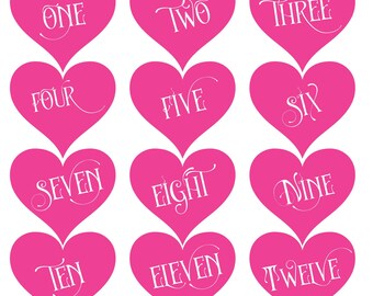May include: Twelve pink heart-shaped cards with white numbers one through twelve written in a decorative script font.