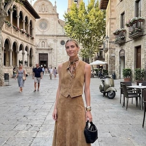May include: A woman in a tan suede vest, midi skirt, and boots stands in a European town square. She carries a black handbag and wears a patterned scarf and gold bracelets. The background features a church, buildings, and a scooter.