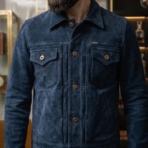 May include: A navy blue suede jacket with a classic design. The jacket features a collared neckline, button-down closure, two chest pockets, and two side pockets. The buttons are a warm, brass colour. The jacket has a tailored fit.