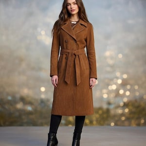 May include: A full-length, brown suede trench coat with a double-breasted design and a matching belt. The coat is worn with black pants and black heeled boots. The background is a blurred mix of neutral tones.