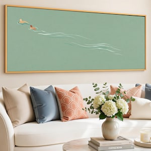 May include: A rectangular framed artwork depicts two figures swimming in a light green body of water. The artwork is framed in a gold-colored frame. The artwork hangs above a white sofa with several decorative pillows in shades of beige, blue, and coral.