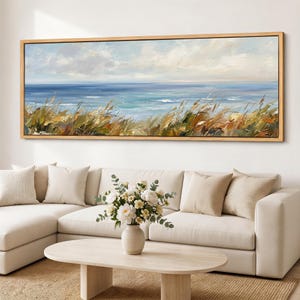 Puede incluir: Una pintura de paisaje panorámico en un marco de madera clara que representa una escena costera con hierbas doradas altas en primer plano, un océano azul tranquilo en el plano medio y un cielo suave y nublado arriba, sobre un sofá beige.