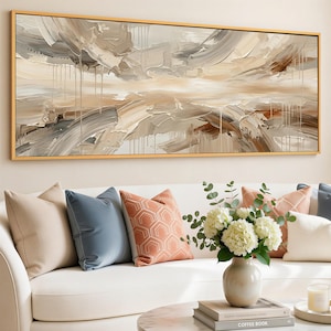 Op de afbeelding: Een abstract schilderij in een gouden lijst, met getextureerde penseelstreken in beige, bruin en grijs. Het kunstwerk hangt boven een witte bank met blauwe en koraalkleurige kussens. Een vaas met witte bloemen staat op een salontafel.