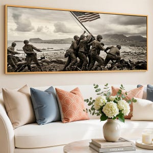 Pode incluir: Fotografia a preto e branco emoldurada que retrata soldados a içar a bandeira americana. A imagem, numa moldura dourada, está pendurada acima de um sofá branco com almofadas decorativas e um vaso de flores.