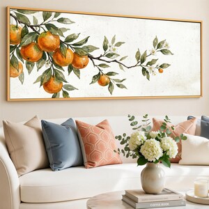 Op de afbeelding: Een aquarel schilderij van een tak met sinaasappels en groene bladeren in een gouden lijst. Het schilderij wordt tentoongesteld boven een witte bank met decoratieve kussens en een vaas met witte bloemen.