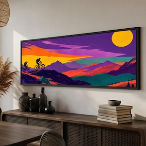 Op de afbeelding: Een rechthoekige wandkunstprint in een zwarte lijst met een gestileerd landschap met twee fietsers in silhouet op een bergkam. De scène toont een levendige zonsondergang met paarse, oranje en gele tinten, een grote gele zon en gelaagde bergketens in tinten van groenblauw, rood en dieppaars.