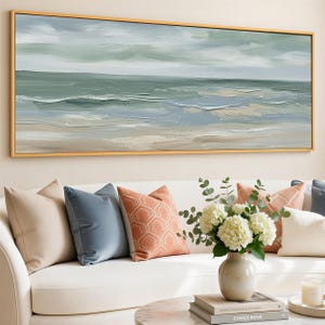 May include: An abstract seascape painting in a gold frame, depicting ocean waves and a cloudy sky in shades of green, blue, and beige. The painting hangs above a white sofa with decorative pillows in beige, blue, and coral colors.