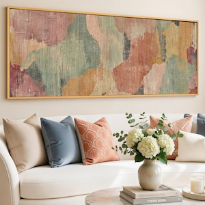 May include: A large rectangular abstract art piece with a gold frame hangs above a white sofa. The artwork features blended colors of pink, green, and orange. Decorative pillows in various colors and patterns adorn the sofa, along with a vase of white flowers.