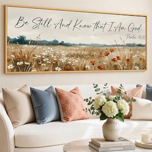 May include: A framed canvas art print with a field of wildflowers and the text "Be Still And Know that I Am God, Psalm 46:10." The artwork features a gold frame and is displayed above a white sofa with decorative pillows.