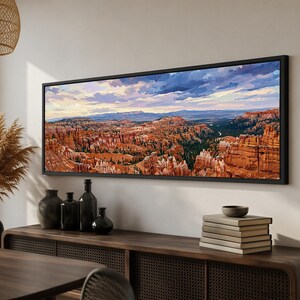 May include: A panoramic landscape painting in a black frame depicting a canyon with orange rock formations, green pine trees, and a cloudy sky with shades of blue, purple, and yellow. The artwork is displayed horizontally on a light wall.
