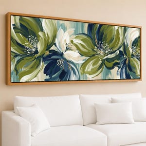 May include: Abstract floral painting in a rectangular wooden frame. The artwork features large, stylized flowers in shades of green, white, and navy blue against a light blue and beige background. The painting is displayed above a white sofa.