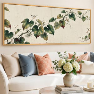 May include: A framed botanical print featuring a vine with large green and red-tinged leaves and small white flowers. The artwork is set against a cream-colored background and framed in gold. The image is displayed above a white sofa with decorative pillows.