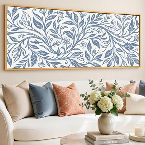 Può includere: Una grande stampa artistica rettangolare su tela con cornice dorata. L'opera presenta un motivo floreale blu e bianco. L'immagine mostra anche un divano bianco con cuscini decorativi di vari colori, un vaso di fiori e un tavolino.