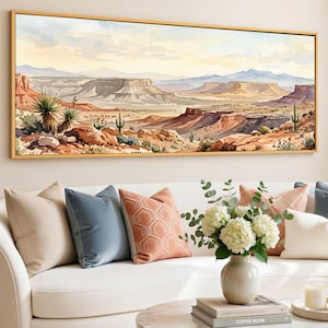 May include: A watercolor painting of a desert landscape with mesas, cacti, and a golden frame. The artwork is displayed above a white sofa with decorative pillows in shades of blue, peach, and beige. A vase of white flowers and a coffee table complete the scene.