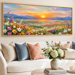 Könnte beinhalten: Ein lebendiges Landschaftsgemälde zeigt einen Sonnenuntergang über einem Feld mit Wildblumen. Das Kunstwerk hat einen goldenen Rahmen und eine Palette warmer Farben, darunter Gelb, Orange und Rosa. Das Gemälde hängt über einem weißen Sofa mit dekorativen Kissen und einer Blumenvase.
