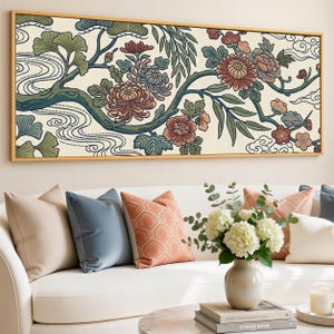 Op de afbeelding: Een ingelijste bloemenkunstprint met een tak met bloemen in tinten bordeauxrood, blauwgroen en groen tegen een crèmekleurige achtergrond. Het kunstwerk, in een gouden lijst, hangt boven een witte bank met decoratieve kussens.