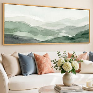 May include: A framed watercolor painting depicts a layered mountain landscape in shades of green and gray. The artwork hangs above a white sofa adorned with decorative pillows in beige, blue, and coral tones. A vase of white flowers and a coffee table complete the scene.