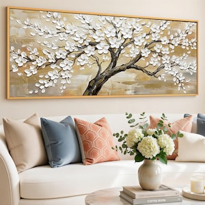 Op de afbeelding: Een ingelijst schilderij van een boom met witte bloesems. Het kunstwerk heeft een bruine stam en takken tegen een beige en gouden achtergrond. Het schilderij wordt tentoongesteld boven een witte bank met decoratieve kussens.