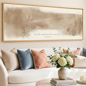 May include: A framed canvas art piece with a gold frame, featuring a neutral abstract design in shades of brown and beige. The text "Be still, and know that I am God. Psalm 46:10" is printed on the canvas. Decorative pillows and a vase of flowers are in the foreground.