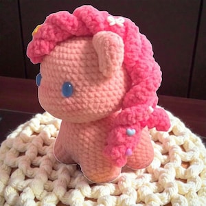 Könnte beinhalten: Ein handgefertigtes Häkelpferd in Rosatönen. Das Pony hat eine flauschige rosa Mähne und einen rosa Schweif, blaue Augen und eine kleine weiße Schleife. Das Pony sitzt auf einer cremefarbenen Häkeloberfläche.