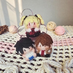 May include: Three handmade crochet keychains featuring cartoon characters. The keychains have different hair colors and outfits. The keychains are attached to a silver metal ring. The background includes yarn balls and a crocheted surface.