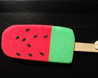May include: A watermelon-shaped sponge with a wooden stick handle. The sponge is red with black seed details on one half and green on the other. A white string is attached to the handle.