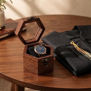 May include: A dark brown wooden hexagonal box with a hinged glass lid and a metal latch, containing a round analog wristwatch with a dark blue face and gold-tone hands. Beside the box lies a black fabric graduation gown with a gold tassel.