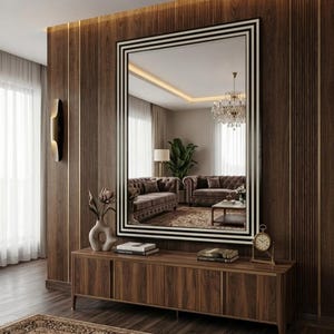 May include: A large rectangular wall mirror with a black and white striped frame, mounted above a long wooden console table. The mirror reflects a living room interior featuring a brown sofa, a chandelier, and a green potted plant.