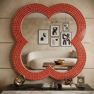 May include: A large, clover-shaped wall mirror featuring a vibrant red mosaic tile frame with a grid pattern. The mirror reflects a room with a cream-colored sofa, a wooden coffee table, and several framed black and white abstract art prints.