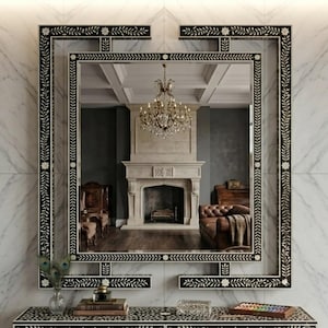 May include: A rectangular wall mirror featuring a black frame with white floral and vine inlay patterns. The mirror reflects a room with a stone fireplace, a crystal chandelier, and a brown leather sofa, creating a layered visual effect.