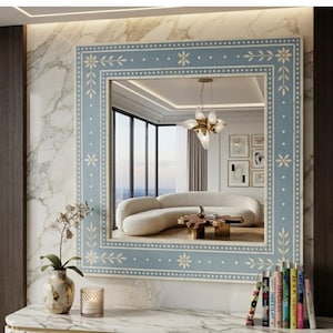 May include: A square wall mirror featuring a light blue frame with white inlaid floral and dotted patterns. The mirror reflects a room with a curved white sofa, a wooden coffee table, and a multi-bulb pendant light fixture hanging from the ceiling.