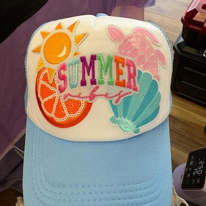 Puede incluir: Una gorra tipo camionero de color azul claro con un panel frontal blanco que presenta parches bordados de un sol amarillo, una rodaja de naranja, una tortuga marina rosa y una concha marina turquesa. El texto "SUMMER vibes" está bordado en el centro.