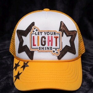 May include: A yellow and white trucker hat with a mesh back. The front features the text "LET YOUR LIGHT SHINE" in a rectangular patch, flanked by brown star-shaped patches. Additional brown stars are embroidered on the brim.