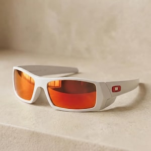 Pode incluir: Óculos de sol brancos com um design de armação envolvente e lentes laranja refletoras. As hastes exibem um pequeno logótipo retangular vermelho na lateral. Os óculos têm uma construção desportiva e elegante.