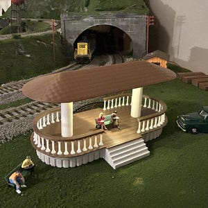 May include: A miniature model train scene featuring a brown gazebo with white pillars and a curved railing. The scene includes a green pickup truck, miniature figures, train tracks, a tunnel, and a yellow train.